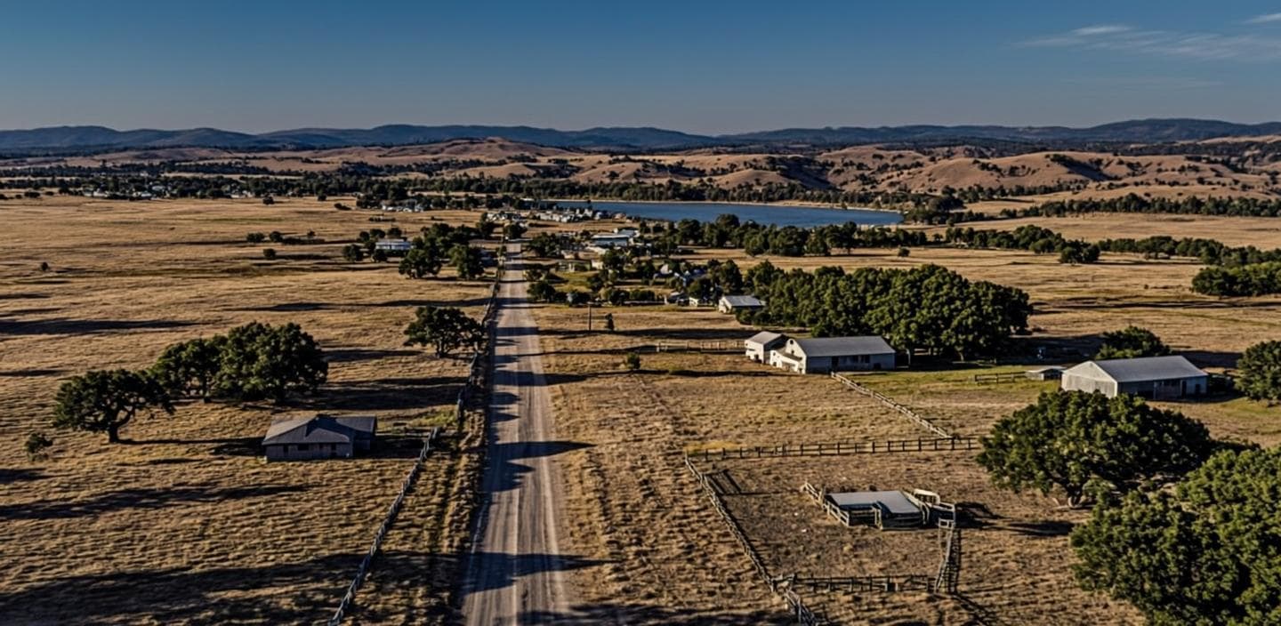 Wide-open ranch country along the SR-79 corridor in Winchester, CA at golden dusk — a long country road, oak-dotted acreage, split-rail fences and a horse paddock, with distant reservoir and new rooftops on the horizon